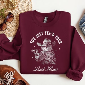 May include: Burgundy sweatshirt with a white graphic of a cat wearing a cowboy hat and holding a gun. The text reads "You just yee'd your last haw".