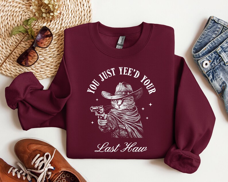 May include: Burgundy sweatshirt with a white graphic of a cat wearing a cowboy hat and holding a gun. The text reads "You just yee'd your last haw".