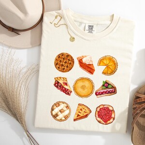 May include: Cream-colored t-shirt featuring watercolor illustrations of various pies. The shirt showcases a collection of baked goods, including whole pies and slices, with lattice crusts and fruit fillings. A hat, sandals, and jewelry are also in the image.