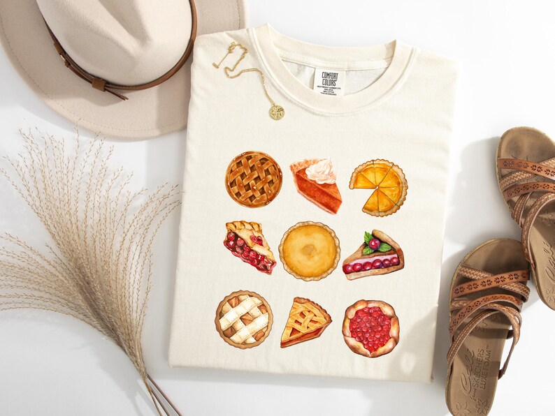 May include: Cream-colored t-shirt featuring watercolor illustrations of various pies. The shirt showcases a collection of baked goods, including whole pies and slices, with lattice crusts and fruit fillings. A hat, sandals, and jewelry are also in the image.