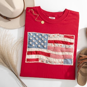 Peut inclure: Un t-shirt rouge avec un motif de drapeau am&eacute;ricain en patchwork. Le drapeau pr&eacute;sente des &eacute;toiles en denim bleu et un tissu &agrave; rayures rouges et blanches. Un collier en or, un chapeau beige et des sandales marron sont &eacute;galement dans l'image.