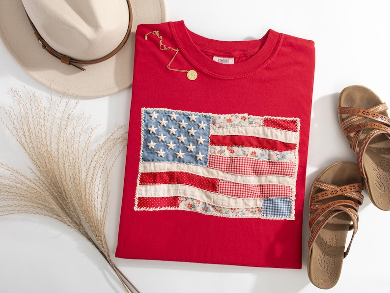 Peut inclure: Un t-shirt rouge avec un motif de drapeau am&eacute;ricain en patchwork. Le drapeau pr&eacute;sente des &eacute;toiles en denim bleu et un tissu &agrave; rayures rouges et blanches. Un collier en or, un chapeau beige et des sandales marron sont &eacute;galement dans l'image.