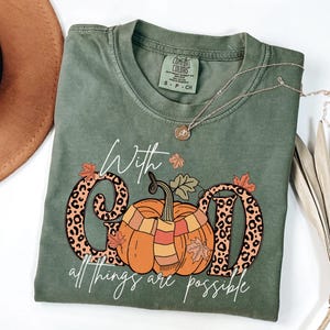 May include: Olive green Comfort Colors t-shirt with a pumpkin graphic and the text "With God all things are possible." The pumpkin has a scarf and is between the letters "G" and "D". A gold necklace is draped across the shirt.