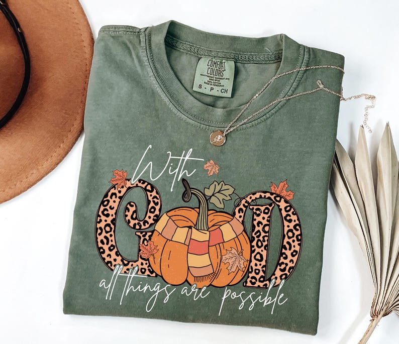 May include: Olive green Comfort Colors t-shirt with a pumpkin graphic and the text "With God all things are possible." The pumpkin has a scarf and is between the letters "G" and "D". A gold necklace is draped across the shirt.