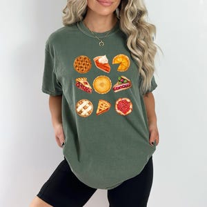May include: Green t-shirt featuring a watercolor illustration of various pies. The design includes whole pies and slices, showcasing different fillings and crusts. The shirt is a comfortable, casual style.