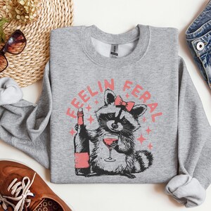 K&ouml;nnte beinhalten: Grauer Pullover mit einem Waschb&auml;ren, der Wein trinkt, und dem Text "Feelin Feral" in Rosa.