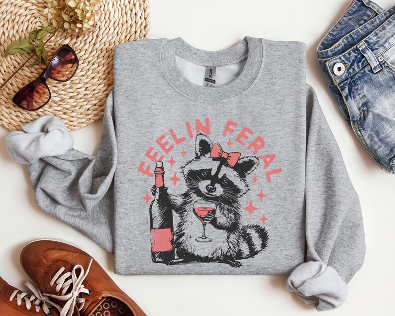 K&ouml;nnte beinhalten: Grauer Pullover mit einem Waschb&auml;ren, der Wein trinkt, und dem Text "Feelin Feral" in Rosa.
