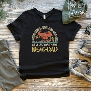 K&ouml;nnte beinhalten: Schwarzes T-Shirt mit einer Vintage-Illustration einer muskul&ouml;sen Comicfigur mit dem Text "Just an ordinary Demi-Dad".