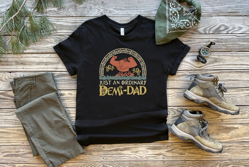 K&ouml;nnte beinhalten: Schwarzes T-Shirt mit einer Vintage-Illustration einer muskul&ouml;sen Comicfigur mit dem Text "Just an ordinary Demi-Dad".