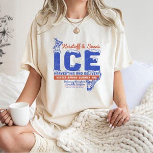 May include: Cream-colored t-shirt featuring a vintage-style graphic with the text "Kristoff & Sven's ICE" in blue, with a snowman and reindeer illustration. The shirt also includes the text "Harvesting and Delivery" and "Winter, Spring, Summer, Fall."
