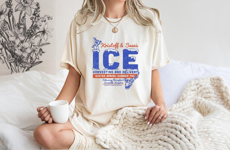 May include: Cream-colored t-shirt featuring a vintage-style graphic with the text "Kristoff & Sven's ICE" in blue, with a snowman and reindeer illustration. The shirt also includes the text "Harvesting and Delivery" and "Winter, Spring, Summer, Fall."