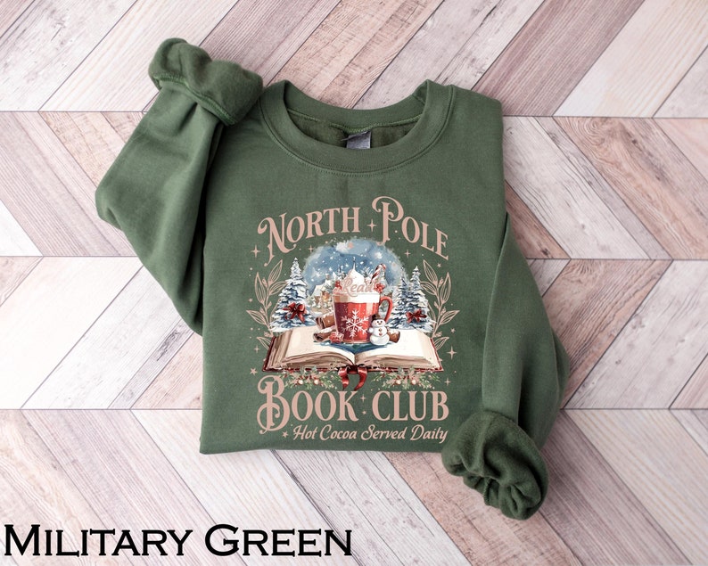 May include: Military green sweatshirt with the text "North Pole Book Club" and a graphic of a snow globe, a mug of hot cocoa, and a snowman. The sweatshirt is a crew neck style.