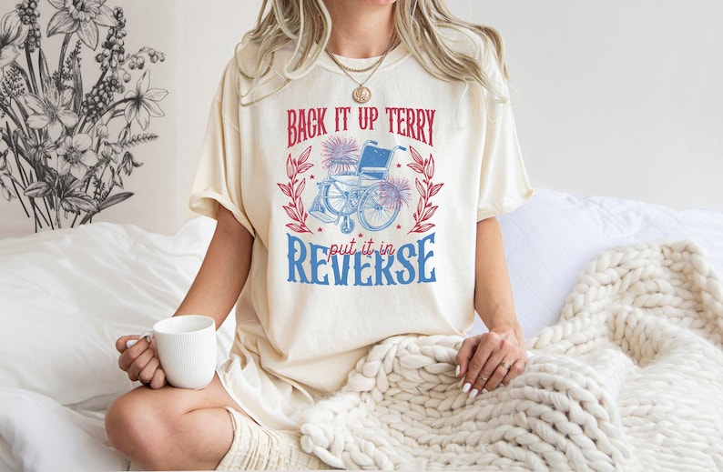 May include: A white t-shirt with a blue and red graphic that says "Back it up Terry put it in reverse". The graphic features a wheelchair with fireworks and a wreath of leaves.