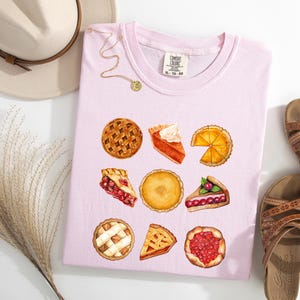 May include: A light pink t-shirt featuring watercolor illustrations of various pies. The shirt is paired with brown sandals, a beige hat, and a gold necklace. The shirt has the text "XL-TG-50".