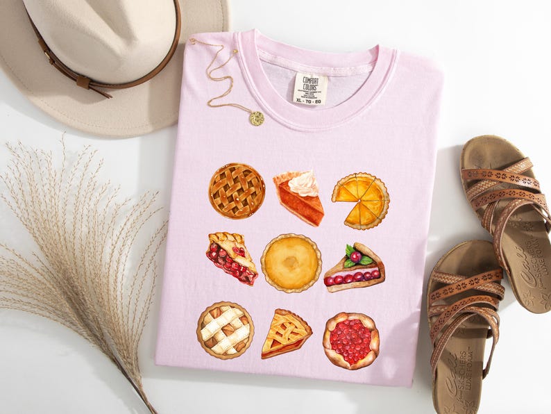 May include: A light pink t-shirt featuring watercolor illustrations of various pies. The shirt is paired with brown sandals, a beige hat, and a gold necklace. The shirt has the text "XL-TG-50".