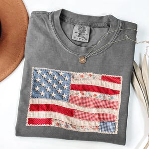 Peut inclure: Un t-shirt gris avec un drapeau am&eacute;ricain cousu. Le drapeau pr&eacute;sente un tissu bleu avec des &eacute;toiles blanches et un tissu &agrave; rayures rouges et blanches. Un collier argent&eacute; avec un petit pendentif repose sur le t-shirt. Le t-shirt est pli&eacute; sur une surface blanche.