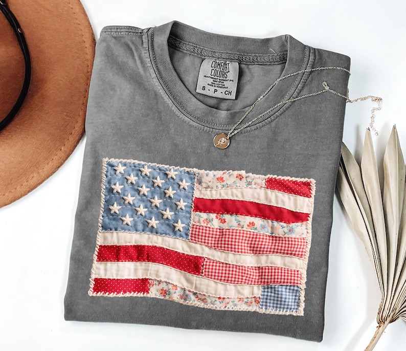 Peut inclure: Un t-shirt gris avec un drapeau am&eacute;ricain cousu. Le drapeau pr&eacute;sente un tissu bleu avec des &eacute;toiles blanches et un tissu &agrave; rayures rouges et blanches. Un collier argent&eacute; avec un petit pendentif repose sur le t-shirt. Le t-shirt est pli&eacute; sur une surface blanche.