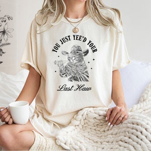 May include: A white t-shirt with a black and white illustration of a cat wearing a cowboy hat and holding a gun. The text on the shirt reads "You just yee'd your last haw".