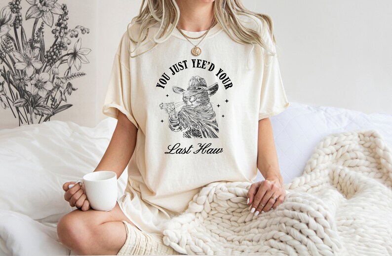 May include: A white t-shirt with a black and white illustration of a cat wearing a cowboy hat and holding a gun. The text on the shirt reads "You just yee'd your last haw".