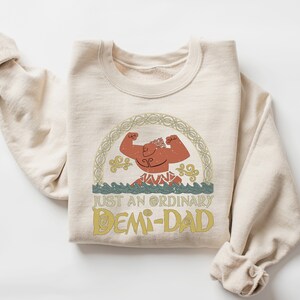 K&ouml;nnte beinhalten: Ein beiger Pullover mit einem Comic-Charakter-Motiv und dem aufgedruckten Text "Just an ordinary Demi-Dad".