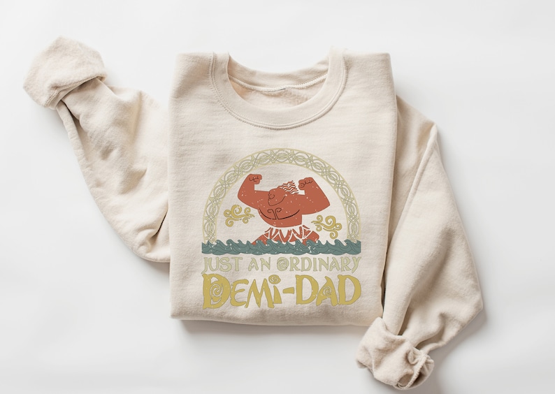 K&ouml;nnte beinhalten: Ein beiger Pullover mit einem Comic-Charakter-Motiv und dem aufgedruckten Text "Just an ordinary Demi-Dad".
