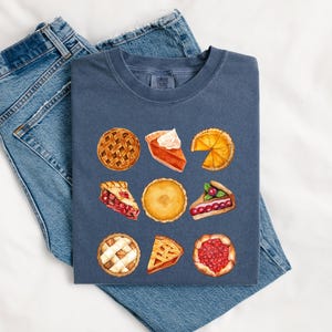 May include: A navy blue t-shirt featuring a watercolor design of various pie slices. The pies include pumpkin, cherry, and lemon, with lattice crusts and whipped cream. The shirt is folded on top of a pair of blue jeans.