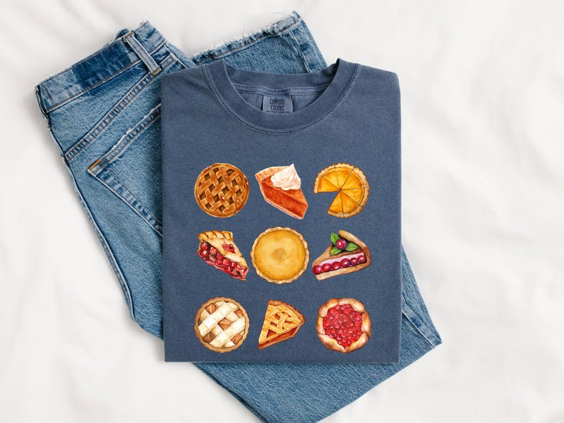 May include: A navy blue t-shirt featuring a watercolor design of various pie slices. The pies include pumpkin, cherry, and lemon, with lattice crusts and whipped cream. The shirt is folded on top of a pair of blue jeans.