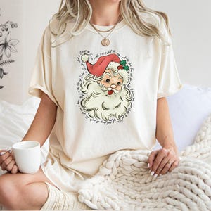 Pu&ograve; includere: T-shirt color crema con grafica vintage di Babbo Natale, con cappello rosso e barba bianca. Il design include testo attorno all'illustrazione di Babbo Natale. Parte di una collezione di abbigliamento a tema natalizio.