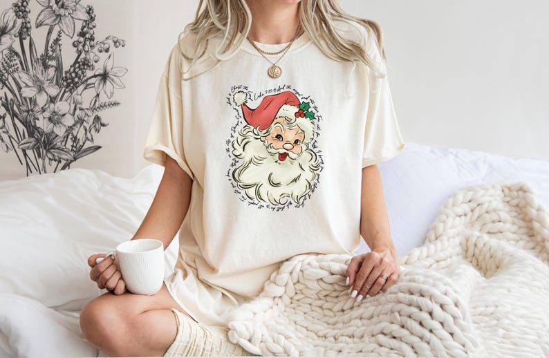 Pu&ograve; includere: T-shirt color crema con grafica vintage di Babbo Natale, con cappello rosso e barba bianca. Il design include testo attorno all'illustrazione di Babbo Natale. Parte di una collezione di abbigliamento a tema natalizio.