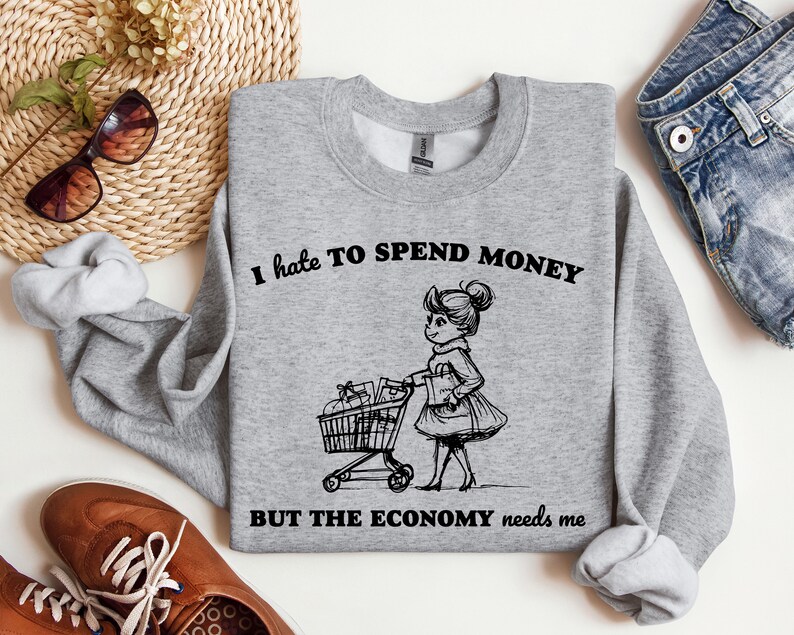 May include: A gray sweatshirt with the text "I hate to spend money but the economy needs me" and a black and white illustration of a person pushing a shopping cart.