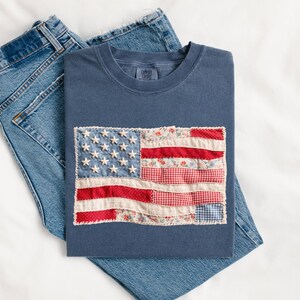 Peut inclure: Un t-shirt bleu marine avec un drapeau am&eacute;ricain en patchwork, pli&eacute; sur un jean bleu. Le drapeau pr&eacute;sente des &eacute;toiles blanches et des rayures rouges et &agrave; motifs floraux. Le t-shirt est en mati&egrave;re souple.