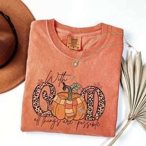 May include: Orange Comfort Colors t-shirt with a pumpkin graphic and the text "With God all things are possible." The pumpkin has a scarf and is surrounded by fall leaves. A gold necklace and a brown hat are also in the image.