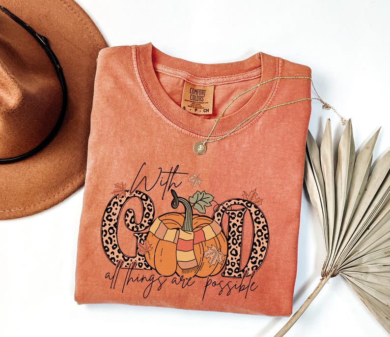 May include: Orange Comfort Colors t-shirt with a pumpkin graphic and the text "With God all things are possible." The pumpkin has a scarf and is surrounded by fall leaves. A gold necklace and a brown hat are also in the image.