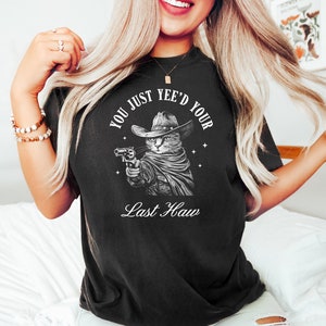 May include: Black t-shirt with a white graphic of a cat wearing a cowboy hat and holding a gun. The text "You Just Yee'd Your Last Haw" is printed below the cat.
