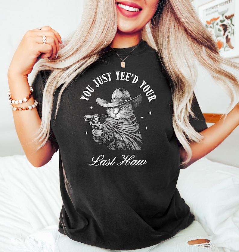 May include: Black t-shirt with a white graphic of a cat wearing a cowboy hat and holding a gun. The text "You Just Yee'd Your Last Haw" is printed below the cat.