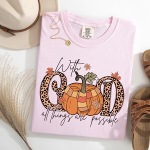 May include: A light pink t-shirt with the phrase "With God all things are possible" and a pumpkin graphic. The letters "G" and "D" have a leopard print design. The shirt is styled with sandals, a hat, and a necklace.