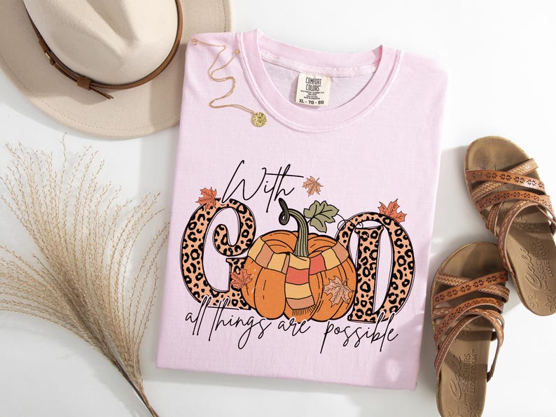 May include: A light pink t-shirt with the phrase "With God all things are possible" and a pumpkin graphic. The letters "G" and "D" have a leopard print design. The shirt is styled with sandals, a hat, and a necklace.