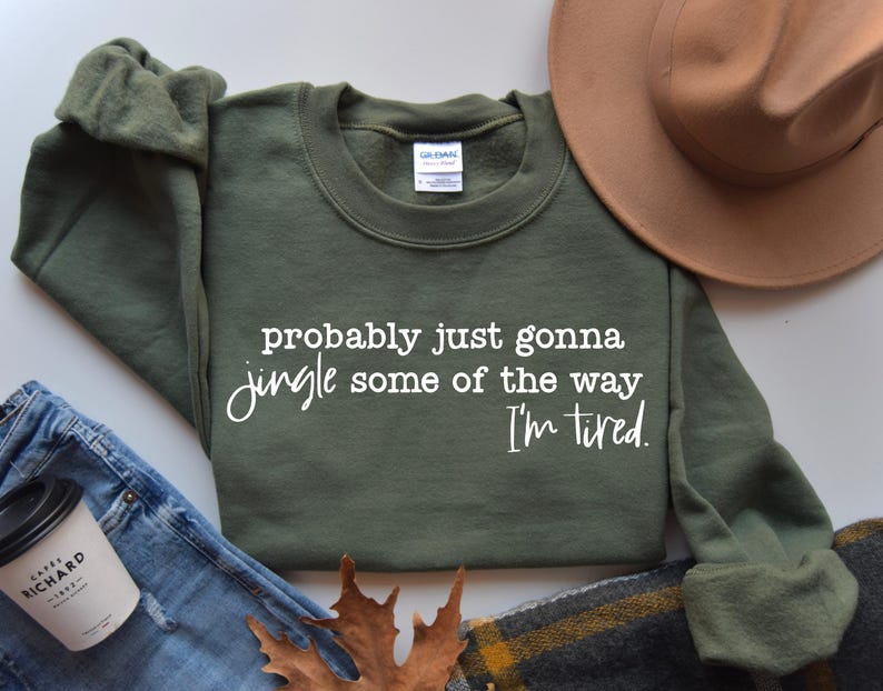 May include: An olive green sweatshirt with the text "probably just gonna jingle some of the way I'm tired" in white script. The sweatshirt is displayed with a brown hat, jeans, and a coffee cup.