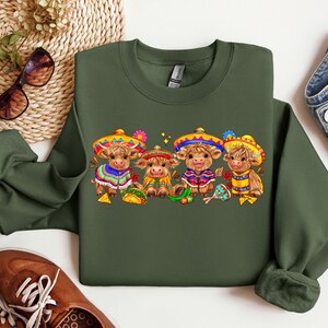 May include: A green sweatshirt with a festive design featuring four brown cows wearing colorful sombreros and holding musical instruments. The cows are surrounded by festive decorations, including flowers, stars, and a maraca.