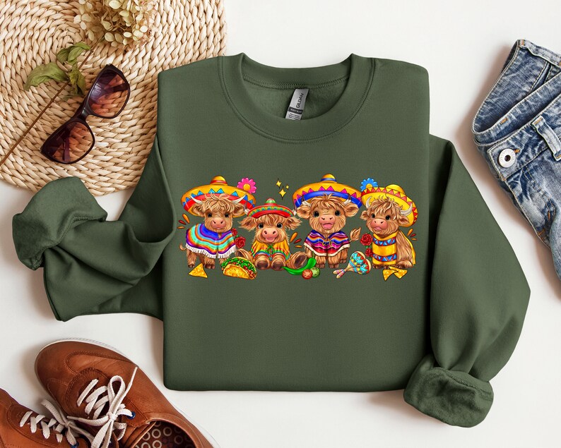 May include: A green sweatshirt with a festive design featuring four brown cows wearing colorful sombreros and holding musical instruments. The cows are surrounded by festive decorations, including flowers, stars, and a maraca.