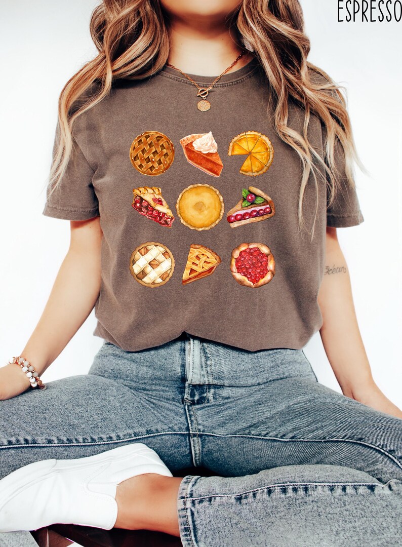 May include: Espresso-colored t-shirt featuring a watercolor illustration of various pies. The design includes whole pies and slices with different fillings, such as cherry, pumpkin, and apple. The shirt is a comfortable, casual style.
