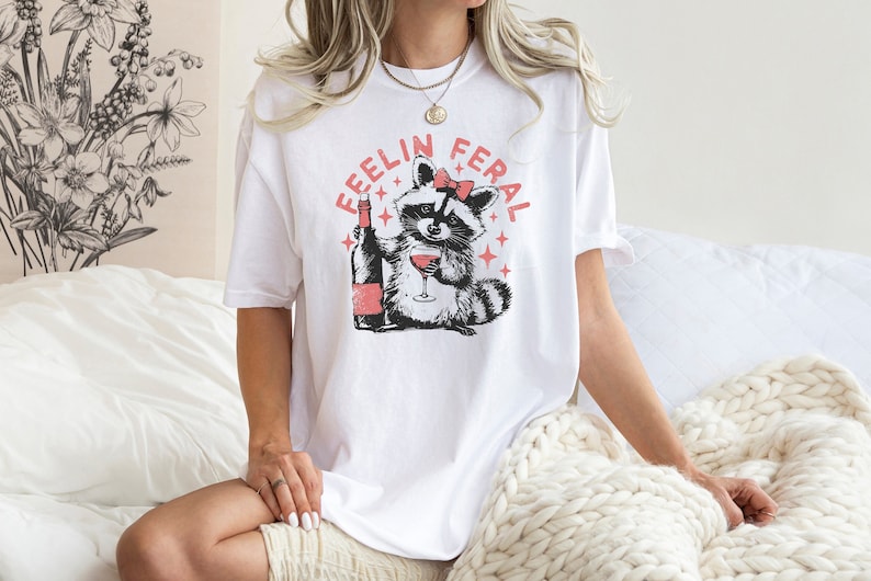 K&ouml;nnte beinhalten: Wei&szlig;es T-Shirt mit einem Grafikdruck eines Waschb&auml;ren, der eine rosa Schleife tr&auml;gt und ein Glas Wein h&auml;lt. Der Text "Feelin Feral" ist in rosa um den Waschb&auml;ren herum geschrieben.