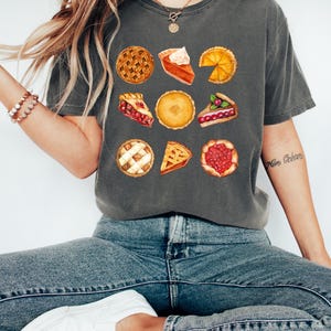 May include: Dark gray t-shirt featuring watercolor illustrations of various pies. The shirt showcases a variety of pie types, including cherry, pumpkin, and lemon, with detailed crusts and fillings. The shirt is paired with blue jeans and white sneakers.