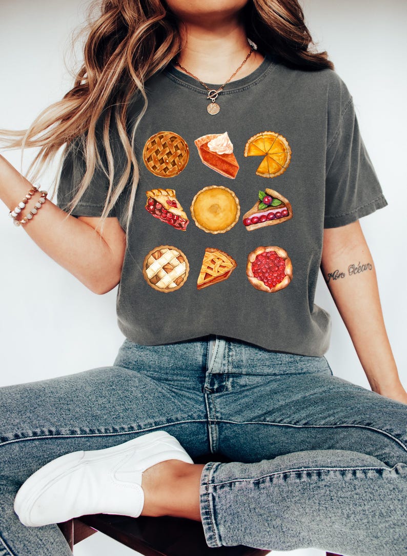 May include: Dark gray t-shirt featuring watercolor illustrations of various pies. The shirt showcases a variety of pie types, including cherry, pumpkin, and lemon, with detailed crusts and fillings. The shirt is paired with blue jeans and white sneakers.