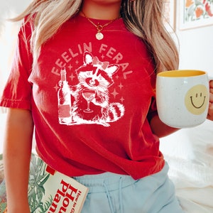 K&ouml;nnte beinhalten: Ein rotes T-Shirt mit dem Text "Feelin Feral" und einem Cartoon-Waschb&auml;r, der Wein trinkt. Der Waschb&auml;r tr&auml;gt eine rosa Schleife. Das Shirt wird von einer Person mit langen blonden Haaren getragen. Die Person h&auml;lt auch ein Buch mit dem Titel "House Plants How to Grow" und eine wei&szlig;e Tasse mit einem gelben Smiley-Gesicht.