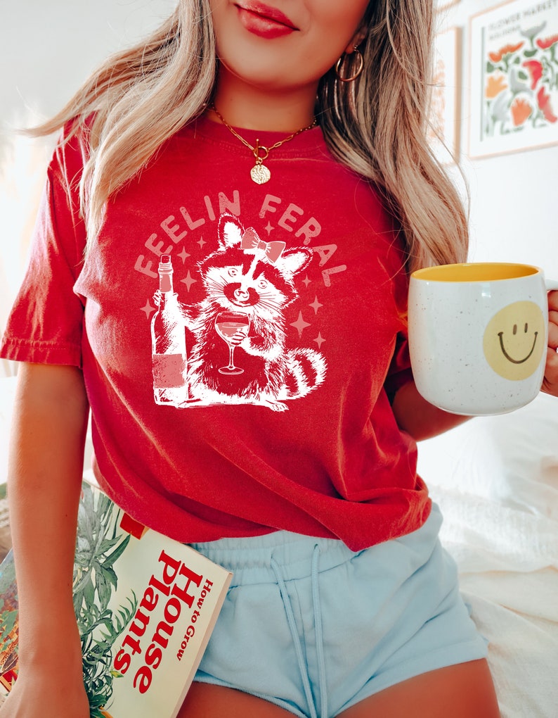 K&ouml;nnte beinhalten: Ein rotes T-Shirt mit dem Text "Feelin Feral" und einem Cartoon-Waschb&auml;r, der Wein trinkt. Der Waschb&auml;r tr&auml;gt eine rosa Schleife. Das Shirt wird von einer Person mit langen blonden Haaren getragen. Die Person h&auml;lt auch ein Buch mit dem Titel "House Plants How to Grow" und eine wei&szlig;e Tasse mit einem gelben Smiley-Gesicht.