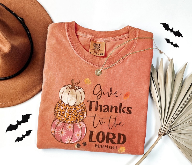 May include: A coral-colored t-shirt with the text "Give Thanks to the LORD" and a stack of pumpkins. The pumpkins have different patterns, including a floral design and a leopard print. A gold necklace and a brown hat are also in the image.