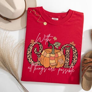 May include: A red t-shirt with a pumpkin graphic and the text "With God all things are possible." The word "GOD" is designed with a leopard print pattern. A gold necklace and sandals are also in the image.