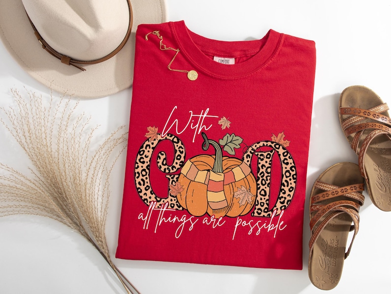 May include: A red t-shirt with a pumpkin graphic and the text "With God all things are possible." The word "GOD" is designed with a leopard print pattern. A gold necklace and sandals are also in the image.