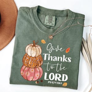 May include: Sage green t-shirt with a stacked pumpkin graphic in cream, leopard print, and pink. The shirt features the text "Give Thanks to the LORD" and "PSALM 136:1". A silver necklace with a pendant is draped across the shirt.